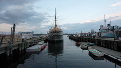 Large cargo ship docked at industrial waterfront with smaller boats and wooden pilings in foreground.