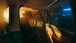 Silhouetted figure wearing a hat inside a vehicle at night, illuminated by warm and cool ambient light.