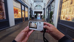 Hands holding a compact digital camera displaying an arcade game on its LCD screen in an upscale shopping passage.