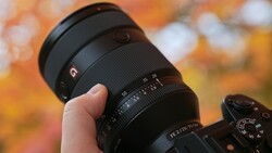 Hand holding a telephoto lens with focus ring and depth-of-field scale visible against blurred autumn foliage background.