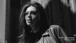 Person wearing clear-framed glasses and light-colored jacket, photographed in black and white with directional side lighting.