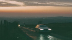 Two cars driving on a wet road at dusk with mountains silhouetted against a twilight sky.
