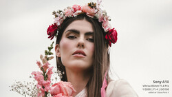 Woman with long dark hair wearing a elaborate floral crown of pink and red flowers with baby's breath, against a neutral background.