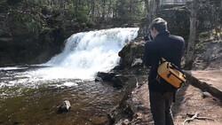 Photographer with camera and yellow bag documents a cascading waterfall in a forested stream setting.
