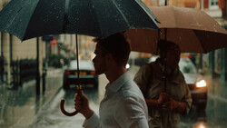 Man holding a dark umbrella in heavy rain on an urban street at dusk.