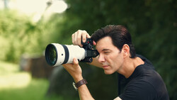 Photographer holding a telephoto lens camera to his eye, positioned in profile against a blurred green outdoor background.