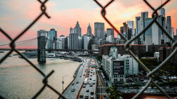 Manhattan skyline at sunset viewed through a chain-link fence from across the water.