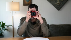 Photographer holding a red digital camera up to his face while seated at a wooden table.