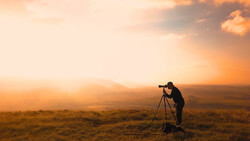 Photographer silhouetted against golden sunset, aiming telephoto lens on tripod across expansive landscape.