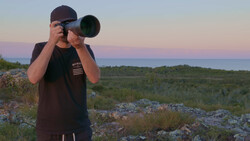 Photographer holding a telephoto lens to his eye while standing on rocky terrain at dusk.