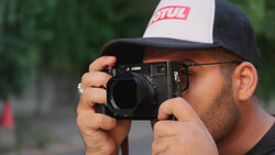 Photographer holding a rangefinder camera up to his face, wearing a trucker-style cap.