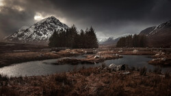 Snow-capped mountain peak rising above forested valley with partially frozen stream in foreground under dramatic cloudy sky.