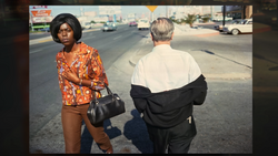 Two people standing on a sunny street with parked cars visible in the background.