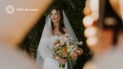 Bride in white dress and veil holding coral and white bouquet in soft-focus garden setting.