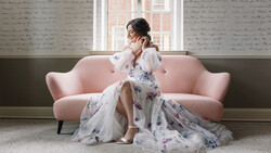 Woman in white blouse and floral skirt sitting on pink sofa near window, touching her face thoughtfully.