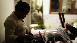 Person working on a weaving loom with colorful yarn threads in soft, directional window light.