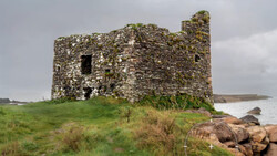 Stone ruins of a multi-story structure with moss-covered walls and empty window openings on a grassy hilltop overlooking water.