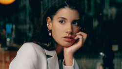 Woman in white blazer resting her chin on her hand, looking toward camera with contemplative expression.