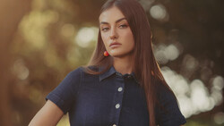 Young woman with long dark hair wearing a navy button-up shirt and red earrings, photographed outdoors with soft golden bokeh background.