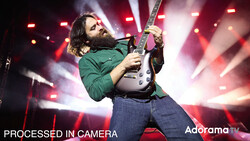 Guitarist performing on stage wearing a green jacket, playing a white electric guitar against red and pink stage lighting.