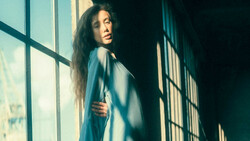 Woman with long dark hair looking over her shoulder by a sunlit window with grid panes.