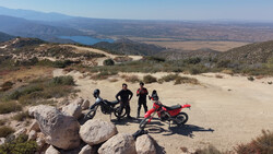 Four people with two motorcycles standing on rocky terrain overlooking a vast landscape with a blue lake in the distance.