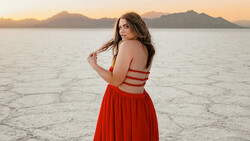 Woman in red strappy dress standing on salt flat at sunset with mountains in background.