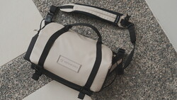 White and black compact camera bag with shoulder strap on gray textured fabric.