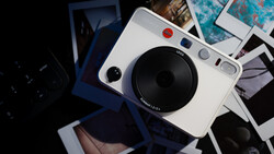 White Leica Sofort instant camera held in hand above scattered instant photographs.