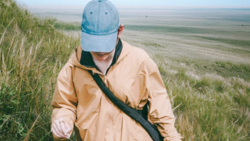Person in tan jacket and denim cap walking through tall grass with expansive plains landscape behind.