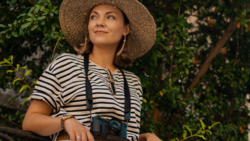 Woman in striped shirt wearing straw hat and holding camera with neck strap in front of green foliage.