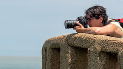 Photographer shooting with a telephoto lens while perched on a concrete structure against a clear sky.