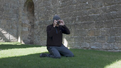Photographer kneeling on grass against stone fortress wall, holding camera to eye to compose a shot.