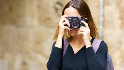 Woman holding a compact mirrorless camera to her face while standing against a textured stone wall.
