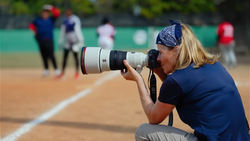 Sports photographer crouching on clay court with telephoto lens during outdoor baseball game.