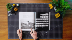 Photographer's hands holding an open book displaying black and white photographs on a grid-patterned cutting mat.