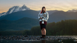Person standing in shallow water holding a camera, with a snow-capped mountain peak in the background at golden hour.