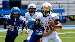 Youth football player in white uniform carrying ball while being tackled by defender in blue uniform.