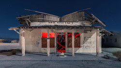 Abandoned roadside diner with boarded red doors and deteriorating wooden roof against clear blue sky.