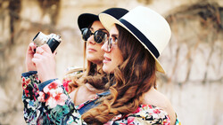 Woman in floral jacket and white Panama hat holding vintage film camera to her face.