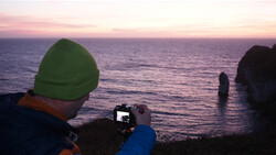 Photographer in green beanie recording sunset seascape on smartphone.