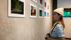 Woman in striped shirt and cream cap viewing framed photographs on gallery wall while holding camera.
