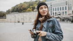 Woman in denim jacket and beanie holding a vintage film camera in an urban plaza.