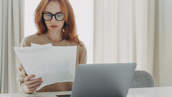 Woman with reddish hair and glasses reviewing documents while working at laptop at desk.