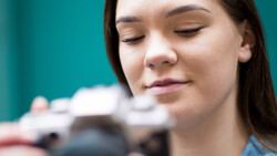 Woman with eyes closed holding a camera, shot against a teal background.