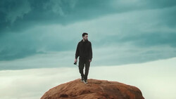 Man in dark clothing standing on top of a large rocky boulder against a dramatic cloudy sky.