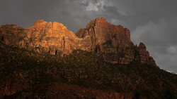 Red rock formations illuminated by golden sunlight beneath a dramatic stormy sky.