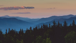 Layered mountain ridges receding into blue-purple haze at sunset, with dark coniferous forest silhouetted in foreground.