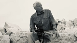 Photographer holding a camera while crouching among rocky terrain in overcast conditions.