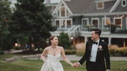 Bride and groom holding hands while walking on manicured lawn in front of large residential home at dusk.